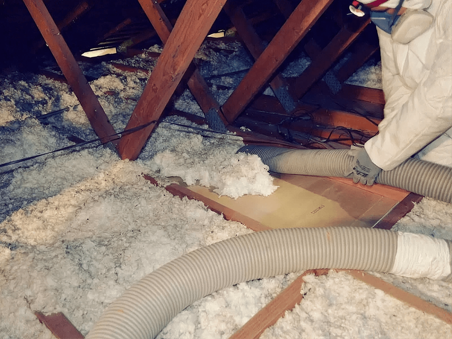 Low-light view inside an attic space showing a worker in a protective suit and mask using a large gray flexible vacuum hose to remove existing loose-fill insulation from between exposed wooden floor joists and roof trusses.