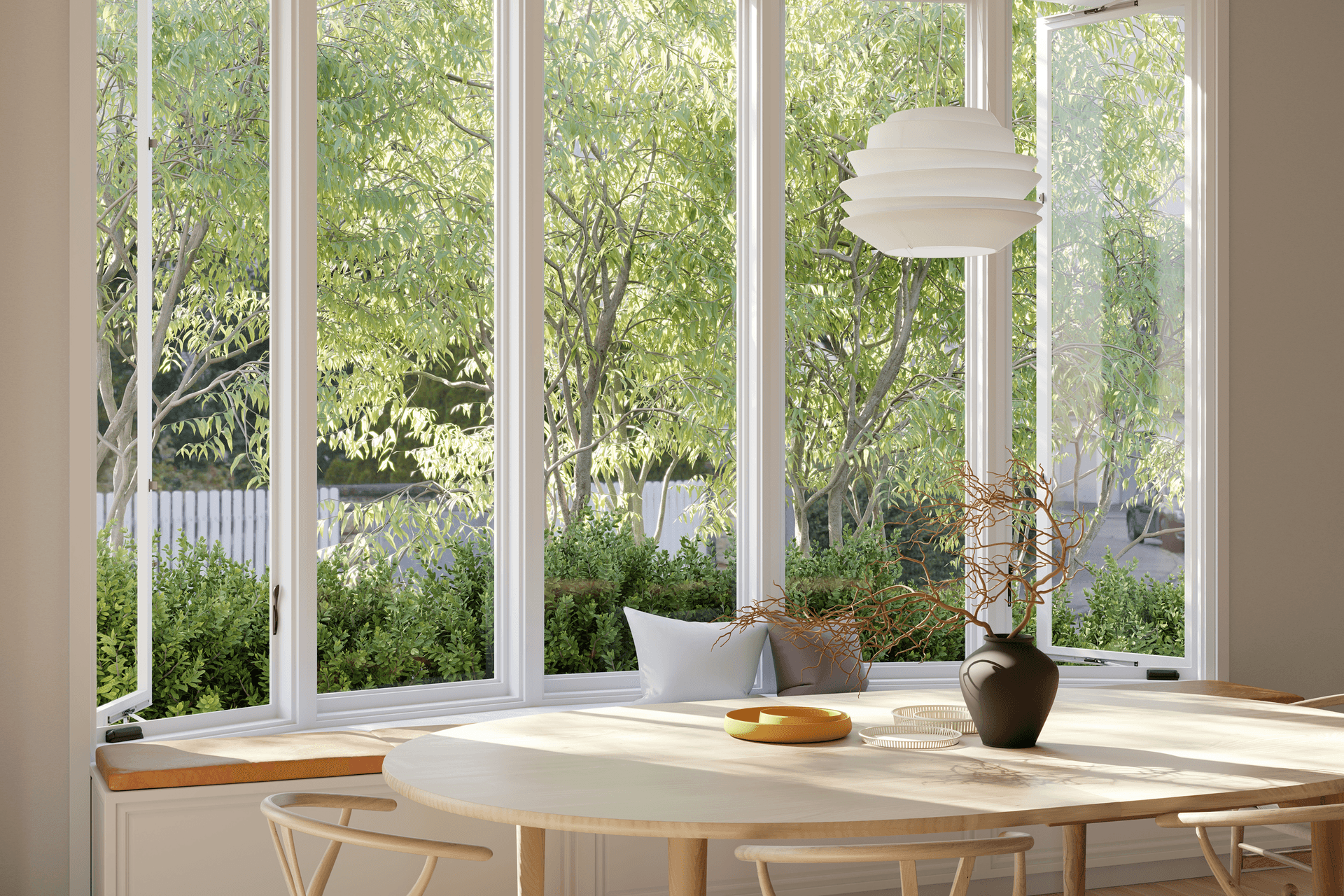 Interior dining room view featuring a large white bow window with side casement panes open to lush green trees, set behind an oval wooden dining table with chairs, a decorative vase, and a modern layered pendant light fixture.