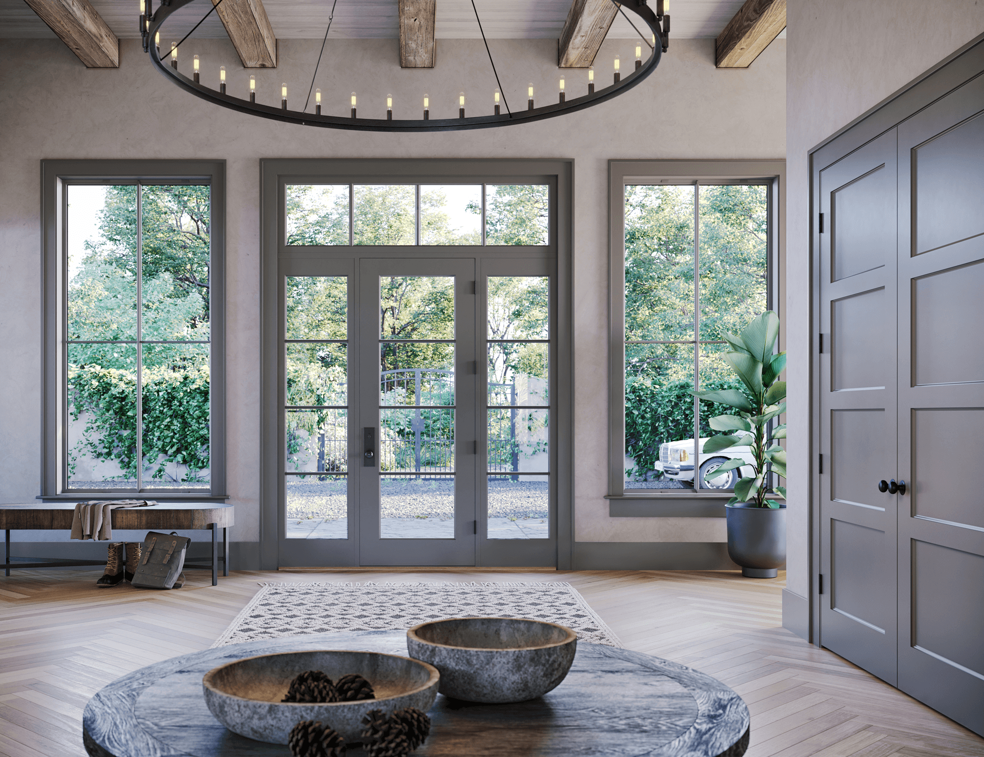 Interior view of a modern farmhouse foyer featuring grey-painted glass-paneled entry doors with matching sidelights and a multi-pane transom window, set alongside tall window units, grey interior doors, exposed ceiling beams, a round wagon-wheel chandelier, and herringbone-patterned wood floors.