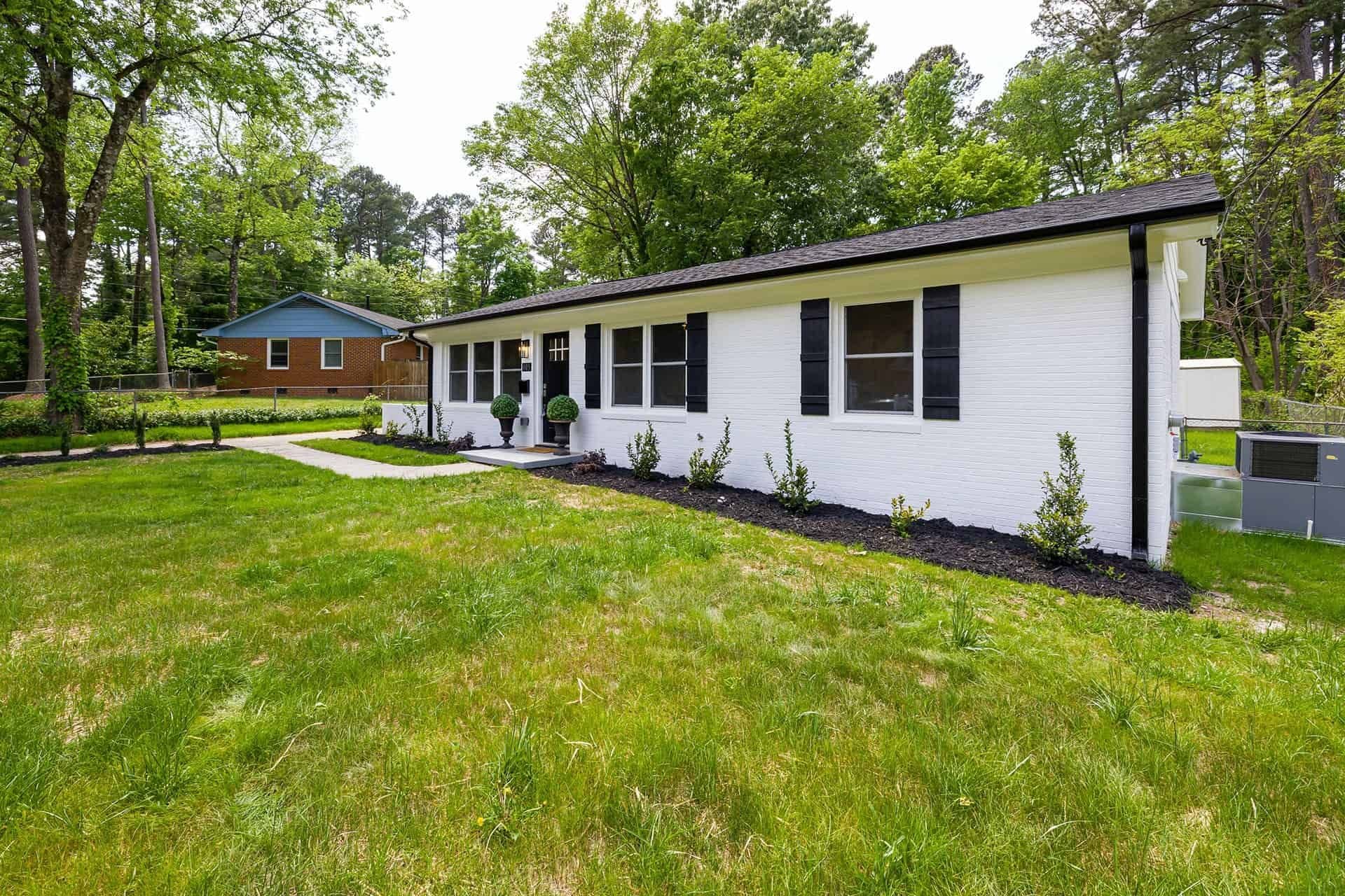 imgi_50_pexels-curtis-adams-8031890.jpg Exterior front view of a single-story white painted-brick ranch home featuring a dark roof, black trim, gutters, and matching black window shutters. The house sits on a green lawn with fresh black mulch landscaping beds along the front foundation and lush green trees in the background.