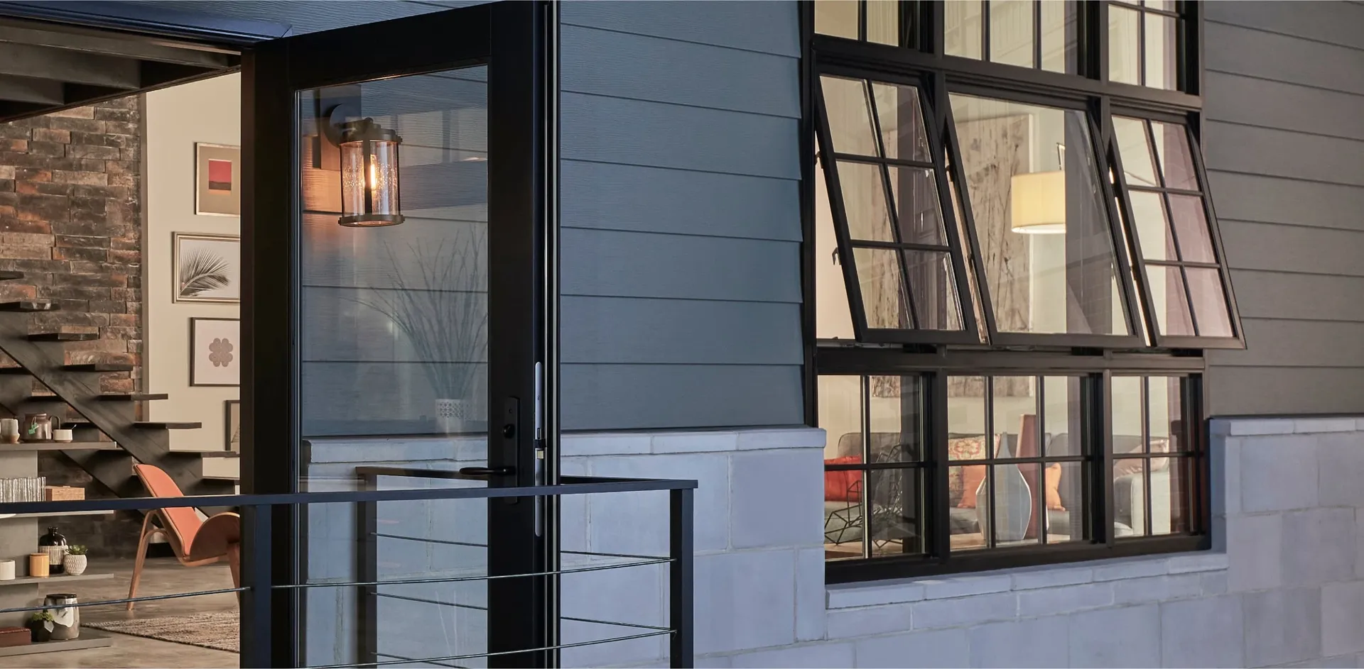 Exterior patio view of a modern house featuring dark grey siding, light grey stone masonry base, a glass door with black trim opening to a balcony railing, and a large black grid window unit with three top-hinged awning panes pushed open for ventilation.