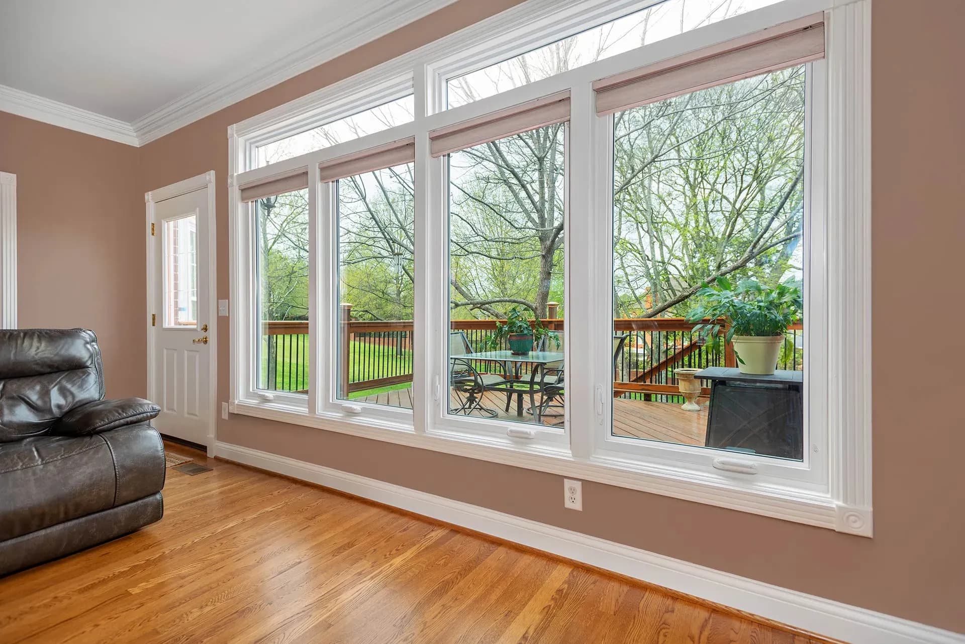 Large sliding glass doors and transom windows opening from living room to wooden deck with outdoor furniture and spring foliage views
