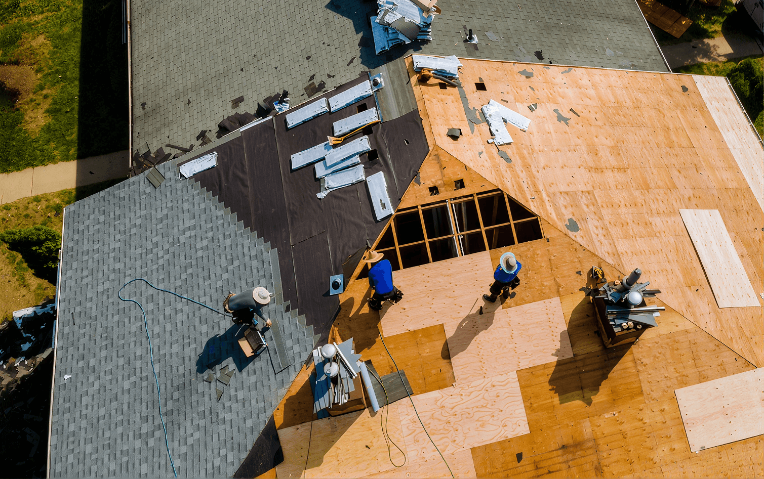 Top-down aerial view of a full roof replacement in progress, showing roofers working across exposed plywood decking, black underlayment, and newly laid grey asphalt shingles.