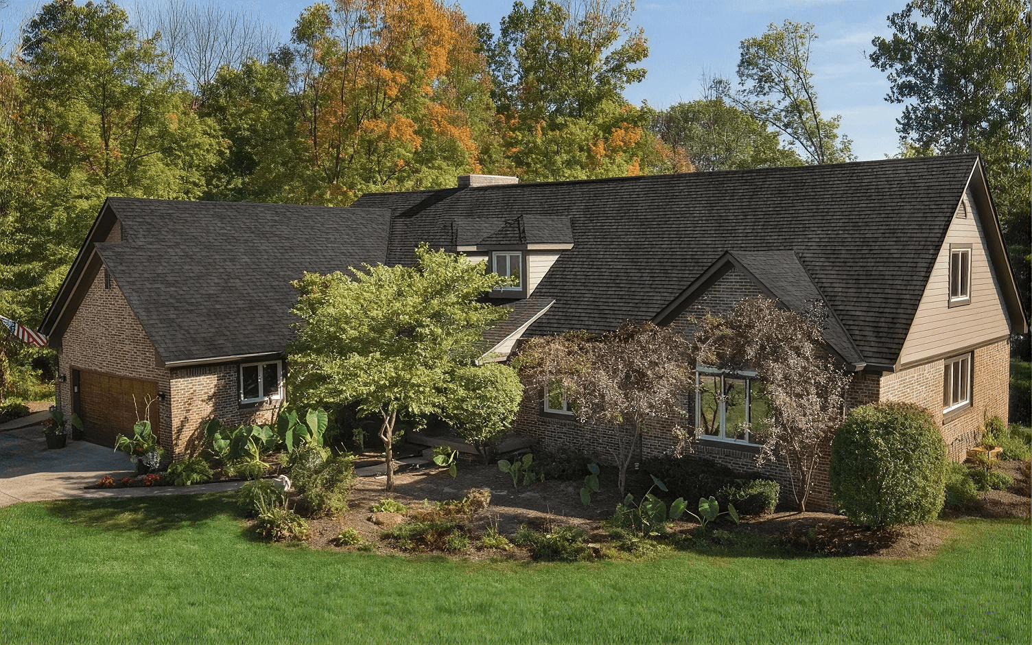 Exterior front view of a large two-story suburban brick house featuring a newly installed dark charcoal asphalt shingle roof, set against a backdrop of autumn trees and a manicured front lawn.