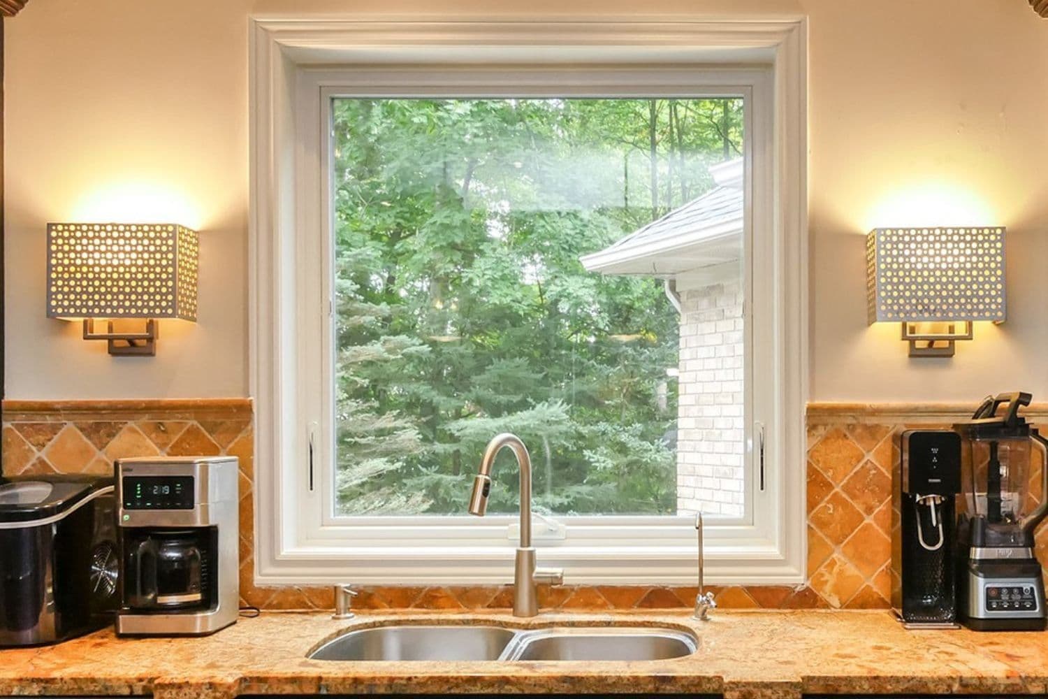 Interior kitchen view featuring a white-framed picture window set above a double-basin stainless steel sink with a gooseneck faucet. The countertop and backsplash consist of warm orange-toned granite and diagonal tile, flanked by small kitchen appliances and two decorative wall sconces on either side of the window framing lush green outdoor foliage.